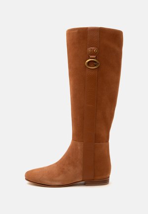 Botte en daim beige montant jusqu’au genou avec une semelle plate et une sangle verticale en cuir dotée d’une boucle ronde en laiton près du haut.