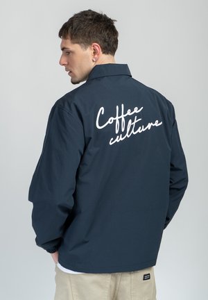 Homme aux cheveux courts et foncés portant une veste bleu marine avec le texte blanc « Coffee culture » dans le dos, pantalon beige, sur fond gris uni.