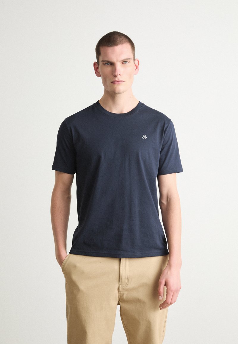 Marineblå bomulds t-shirt med korteærmer, rund halsudskæring og lille broderet logo på brystet; parret med beige bukser.
