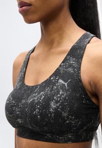 Κοντινό πλάνο ενός ατόμου που φοράει μαύρο και γκρι αθλητικό σουτιέν με σχέδιο και λογότυπο Puma στο αριστερό στήθος, μπροστά σε ένα απλό φωτεινό φόντο.