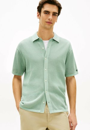 Młody mężczyzna w jasnozielonej, teksturowanej koszuli na krótki rękaw zapinanej na guziki, założonej na białą koszulkę, oraz beżowych spodniach, stojący z rękami w kieszeniach.