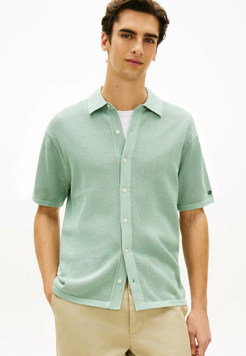 Jeune homme portant une chemise à manches courtes boutonnée texturée vert clair par-dessus un t-shirt blanc et un pantalon beige, debout avec les mains dans les poches.