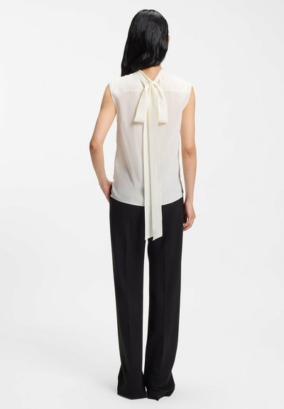 Blusa sin mangas de color crema con un detalle de lazo anudado en la parte trasera, combinada con pantalones negros. Textura suave, tela ligera.