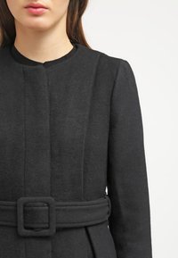 Schwarzer Wollmantel mit rundem Ausschnitt, tailliertem Bund und vertikalen Nähdetails. Glatte Textur, maßgeschneiderte Passform und keine sichtbaren Verschlüsse.