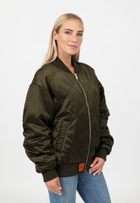 Giacca bomber trapuntata verde oliva con zip frontale, polsini a costine e un'etichetta visibile all'orlo. Materiale lucido e strutturato.