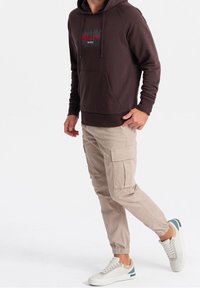 Brązowa bluza z kapturem z przednią kieszenią i czerwonym napisem 'BKLYN', w połączeniu z beżowymi spodniami typu cargo z bocznymi kieszeniami oraz białymi trampkami z niebieskimi akcentami.