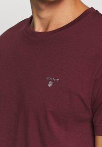 Burgundowa bawełniana koszulka z okrągłym dekoltem, krótkimi rękawami i małym haftowanym logo GANT na piersi. Gładka tekstura.