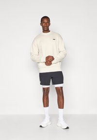 Sweatshirt beige avec des poignets et un ourlet côtelés, porté avec un short gris foncé et des chaussettes de sport blanches, assorti à des baskets blanches.