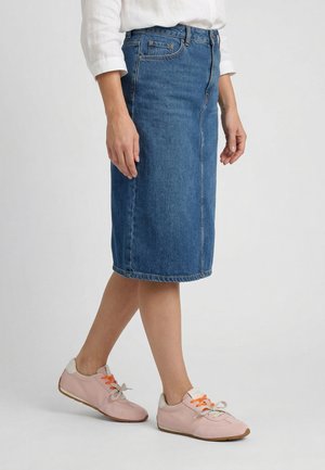 Kvinde iført blå denimnederdel til knæet, hvid skjorte med op-rullede ærmer og pink sneakers med orange snørebånd, går på ensfarvet baggrund.