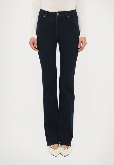 Jean flare bleu marine en tissu denim, doté d'une taille mi-haute, de poches avant et d'une fermeture à boutons, stylisé avec des chaussures blanches à bouts pointus.