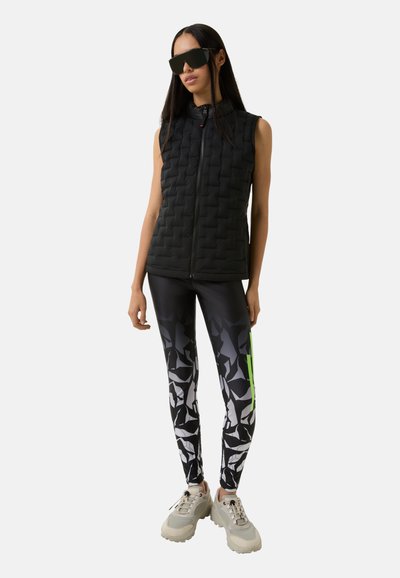 Schwarze, gesteppten ärmellosen Weste mit hohem Kragen, kombiniert mit gemusterten Leggings in Schwarz und Weiß mit grünen Akzenten. Klobige graue Sneakers.