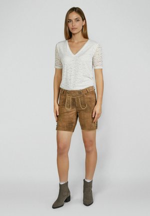 Vrouw draagt een wit kanten top met korte mouwen, bruine suède traditionele korte broek met klep en grijze enkellaarsjes, tegen een effen achtergrond.