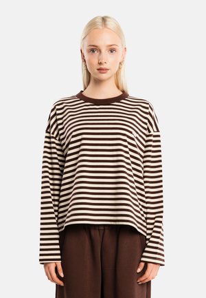 Joven mujer con cabello rubio lleva una camisa de manga larga a rayas marrones y beige y pantalones marrones sueltos, mirando hacia adelante sobre un fondo simple.
