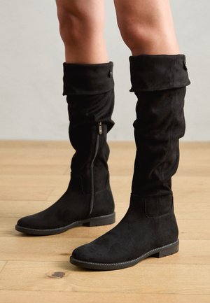 Bottes montantes en daim noir avec un talon bas et une fermeture éclair latérale, portées sur des jambes nues, posées sur un sol en bois.