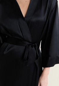 Peignoir enveloppant en satin noir à manches larges, noué en un nœud à la taille, montrant le haut du corps et un bras partiellement d'une personne.