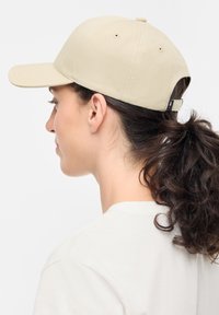 Beige pet met een gebogen rand, stoffen textuur, verstelbare band aan de achterkant en ventilatiegaatjes. Draagt een wit t-shirt.