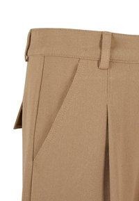 Pantalon en tissu beige avec plis devant, poche latérale inclinée, passants pour ceinture et détail de rabat latéral pour plus de style.