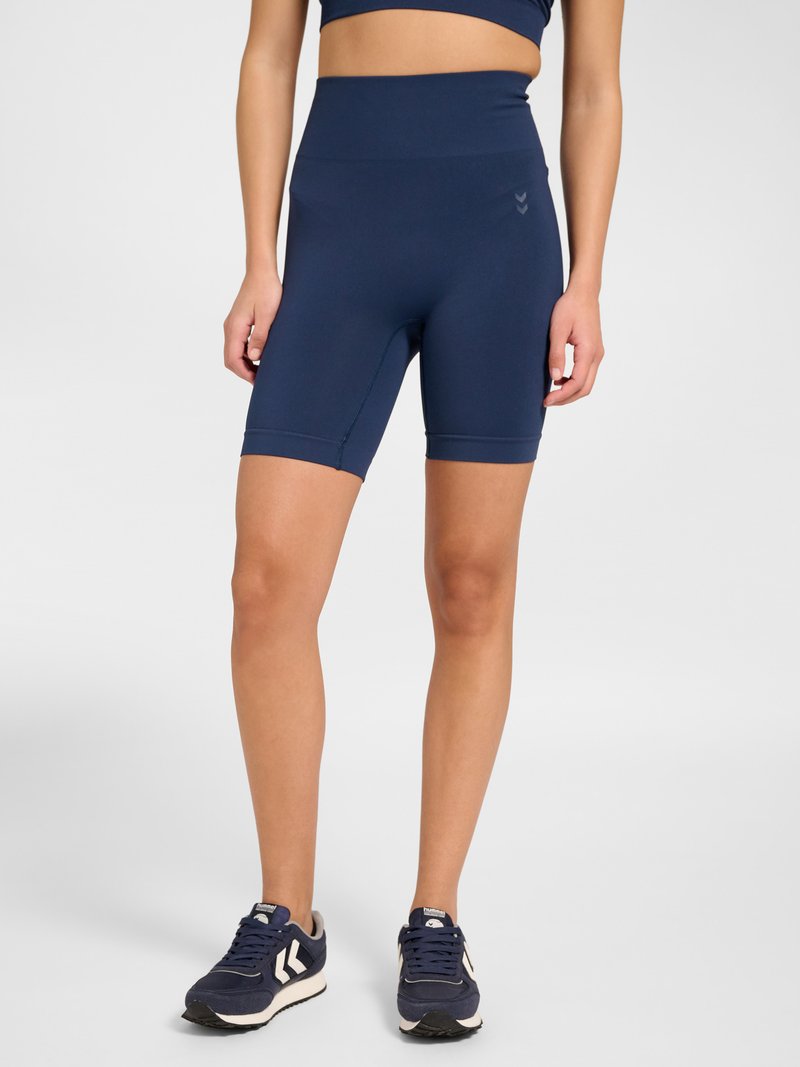 Woman wearing navy blue high-waisted biker shorts and matching crop top with navy blue and white sneakers, standing against a plain background.