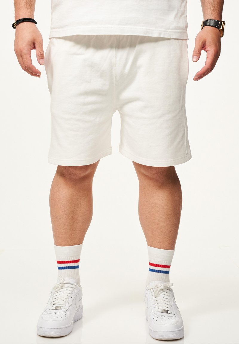 Weiße Baumwollshorts mit elastischem Bund, kombiniert mit weißen Sneakers und Kniestrümpfen mit roten und blauen Streifen.