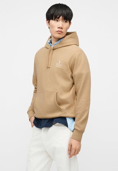 Νεαρός άνδρας που φοράει μια μπεζ φούτερ Polo Ralph Lauren πάνω από στρωμένα μπλε πουκάμισα και λευκό παντελόνι, κοιτάζοντας στο πλάι με τα χέρια στις τσέπες.