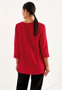 Rød tekstureret pullover med trekvartærmer, der har et perforeret mønster, rund halsudskæring og en afslappet pasform.