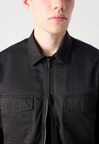 Black lightweight jacket made of smooth fabric, featuring a full zipper, pointed collar, and two chest pockets with flap closures.