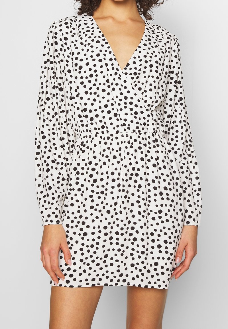 Robe à pois noir et blanc avec un décolleté en V profond, des manches longues, une taille froncée et une coupe décontractée au bas. Texture de tissu lisse.