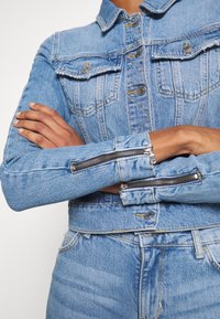Světle modrá džínová bunda s předními kapsami, stříbrným kováním, zipy na manžetách a žlutým prošíváním. Přiléhavý střih zvýrazňuje postavu.