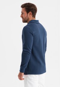 Polo de manga larga azul marino con cuello, que presenta una textura suave y un diseño ajustado. Combinado con pantalones blancos.