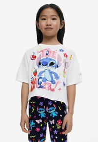 Witte cropped t-shirt met een kleurrijke afbeelding van een blauwe alien en verschillende kleine opdrukontwerpen. Gecombineerd met marineblauwe leggings versierd met bloemen- en karakterpatronen.