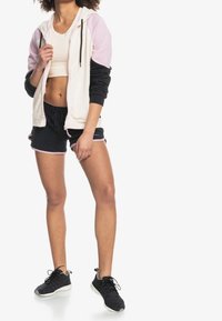 Conjunto deportivo que incluye una sudadera con cremallera color crema con detalles en negro y rosa, combinada con pantalones cortos negros y zapatillas negras.