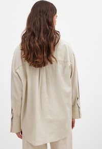 Camicia in lino beige chiaro con una vestibilità rilassata, dotata di maniche lunghe, una piega sulla schiena e polsini con bottoni. Texture morbida e design minimale.
