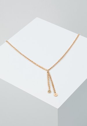 Collier chaîne dorée avec deux petits pendentifs ronds, présenté drapé sur un piédestal géométrique blanc sur un fond gris clair.
