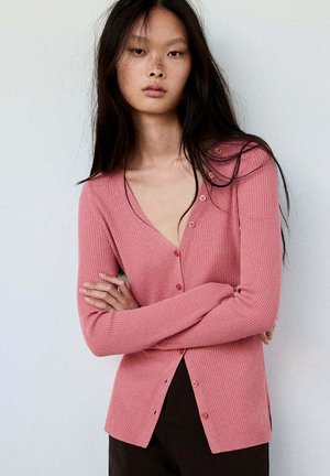 Mujer con cabello largo y oscuro, vestida con un cárdigan rosa de canalé con botones y pantalones negros, de pie con los brazos cruzados contra una pared blanca.