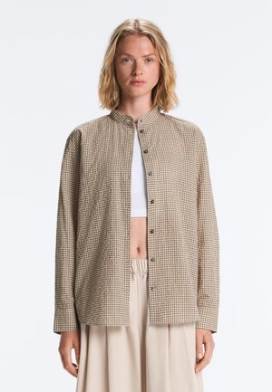 Kvinde med skulderlangt lyst hår iført en beige ternet skjorte over en hvid crop top og løse beige bukser.