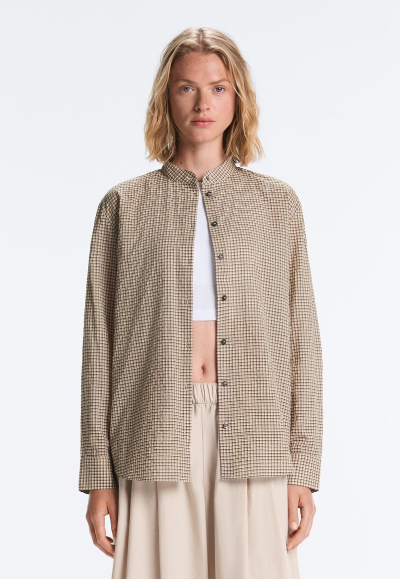 Femme aux cheveux blonds mi-longs portant une chemise beige à carreaux boutonnée sur un crop top blanc et un pantalon beige ample.