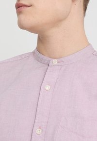 Hellrosa Leinenhemd mit Stehkragen, einer Brusttasche und zwei Knöpfen. Die Textur wirkt glatt und leicht zerknittert.