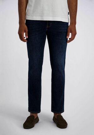 Man draagt donkerblauwe rechttoe-rechtaan spijkerbroek, wit gestructureerd kort shirt met korte mouwen en donkerbruine instapschoenen, staand op een lichte achtergrond.