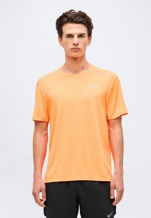 Fiatal férfi áll, előre néz, narancssárga Nike rövid ujjú sportfelsőt és fekete rövidnadrágot visel egyszínű háttér előtt.