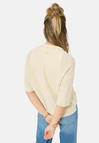 Pull beige clair à manches courtes, avec un motif tricoté texturé et un logo discret dans le dos, associé à un jean bleu clair ample.