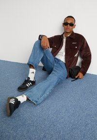 Pruunid kootud jope, valge särk, helesinised teksapüksid, mustad Adidas spordialused valgete triipudega ja väike must kott.