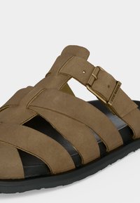 Καφέ σανδάλι από ελαφρώς σουέτ δέρμα με φαρδιές σταυρωτές λουρίδες, ρυθμιζόμενο μεταλλικό πόρπη, μαύρη σόλα και το "Steve Madden" χαραγμένο στην πόρπη.