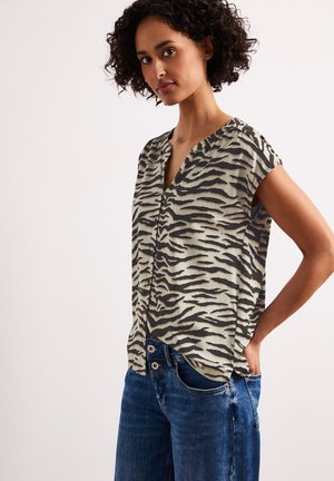 Kobieta w bezrękawniku w nadruk zebry i niebieskich dżinsach, stojąca z jedną ręką w kieszeni na jednolitym tle.