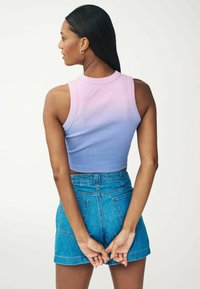 Crop top s gradientem od růžové po modrou, žebrovaná textilie. Nosený s vysokými džínovými šortkami. Modelka otočená zády, s rukama zkříženýma za zády.