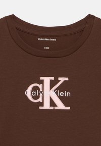 Hnědé bavlněné tričko s kulatým výstřihem a vyšívaným logem "Calvin Klein" v světle růžové a bílé barvě. Velikostní štítek: 8 (128).