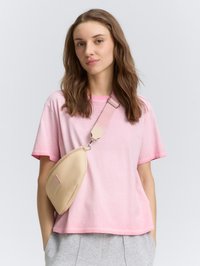 Beige schoudertas van zacht materiaal, met een gebogen vorm, lichte stiksel details en een afneembare schouderband. Gecombineerd met een roze T-shirt.