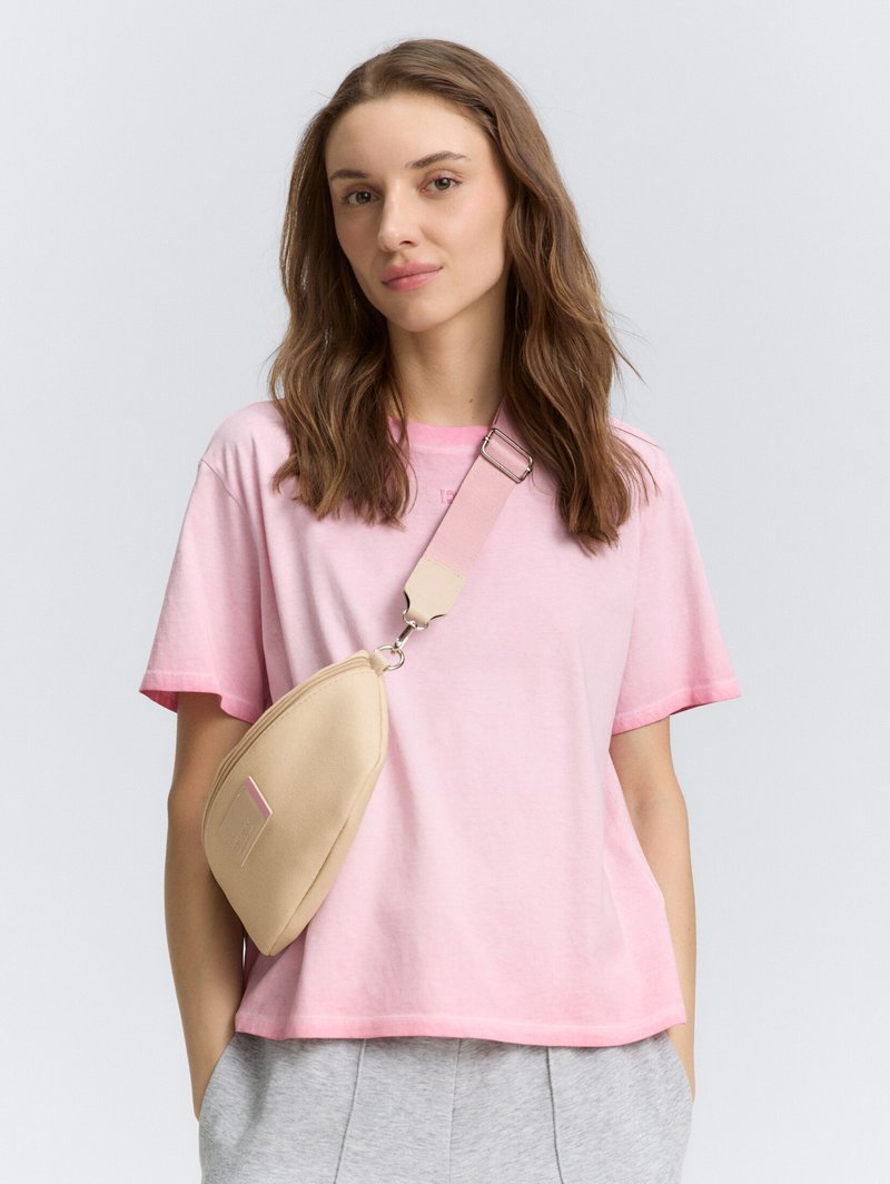 Beige schoudertas van zacht materiaal, met een gebogen vorm, lichte stiksel details en een afneembare schouderband. Gecombineerd met een roze T-shirt.