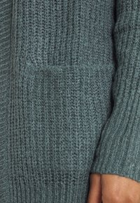 Texturovaný zelený pletený kardigán s vertikálním žebrovaným vzorem, s přední kapsou na levé straně a měkkým, pohodlným materiálem.