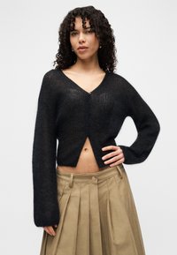 Μαύρη πλεκτή ζακέτα με V-λαιμόκοψη, άνετη εφαρμογή και μακριά μανίκια. Διαθέτει κλείσιμο με κουμπιά και κοντό τελείωμα. Φοριέται πάνω από μια μπεζ πλισέ φούστα.