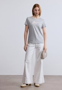 Γκρι κοντομάνικη μπλούζα με τη λέξη "Boss", συνδυασμένη με άσπρο παντελόνι φαρδύ και κρεμ αθλητικά παπούτσια, κρατώντας μια τσάντα ανοιχτό γκρι.